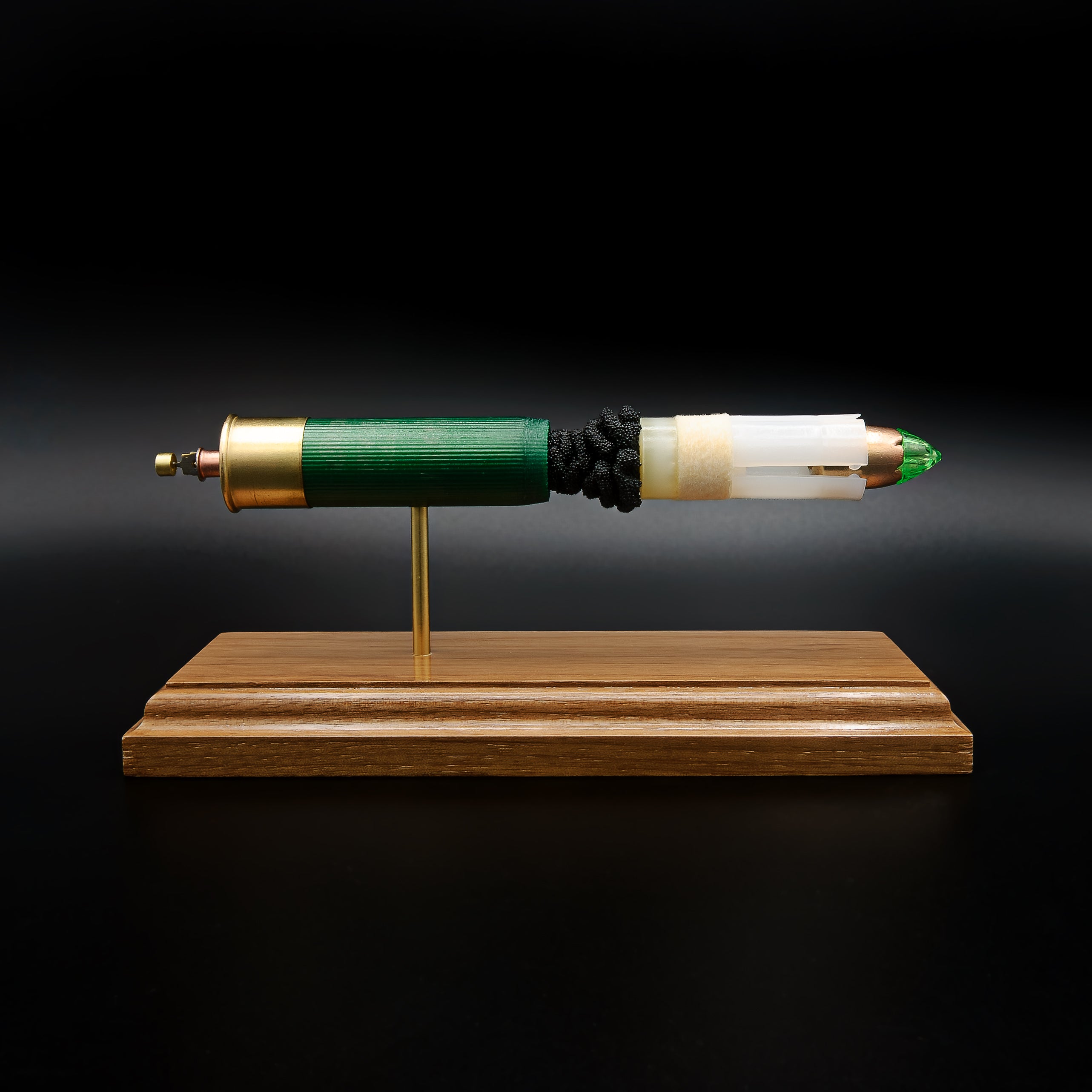 Front profile of an exploded Remington 12 Gauge AccuTip slug display featuring the green hull, exposed simulated powder, sabot, copper slug with green polymer tip, and expanded primer assembly with primer cup, anvil, and battery cup mounted on a handcrafted walnut wood presentation base.