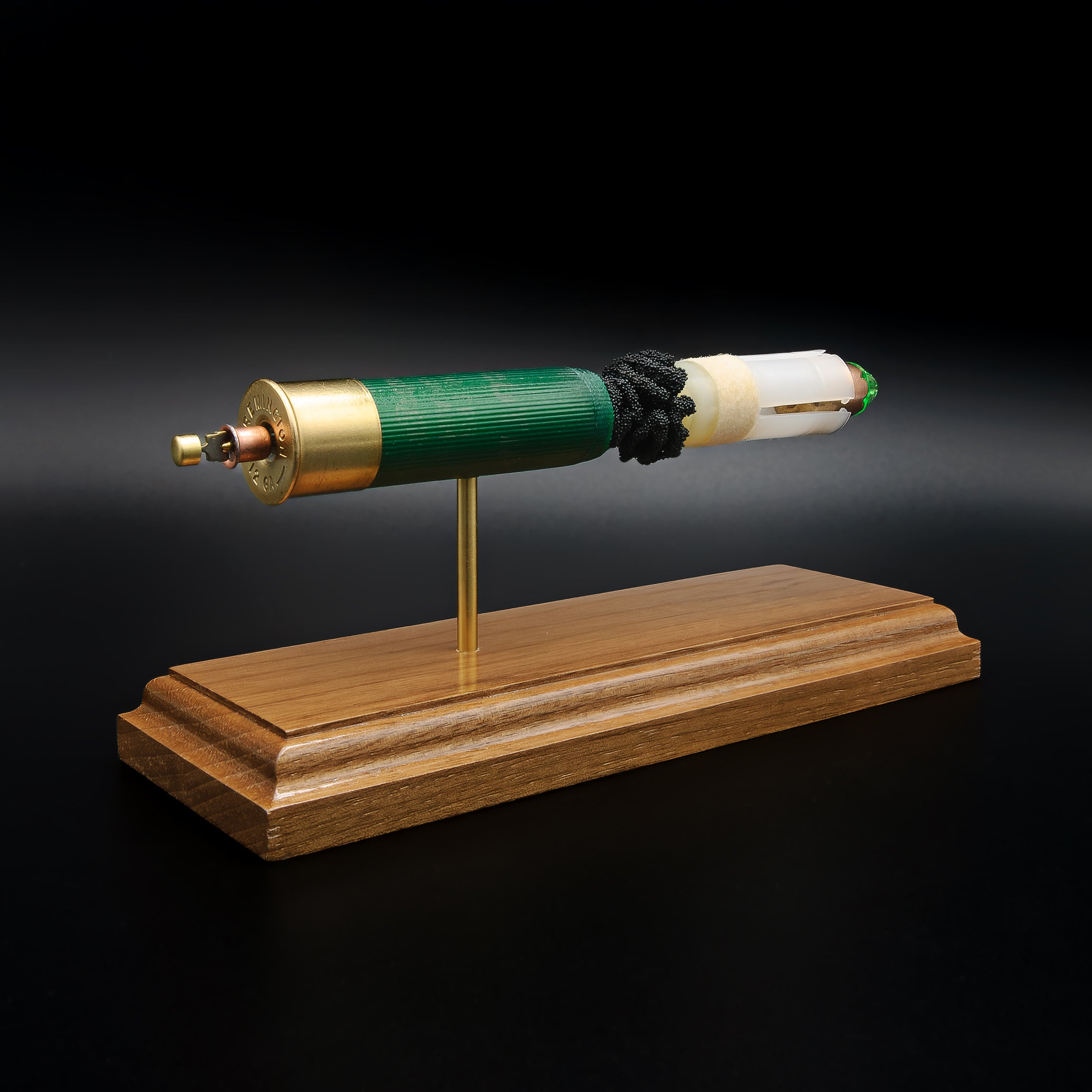 Rear-left three-quarter view of an exploded Remington 12 Gauge AccuTip display emphasizing the brass headstamp, expanded primer cup, anvil, battery cup, green hull, simulated powder, sabot, and copper slug with green polymer tip on a walnut presentation base.