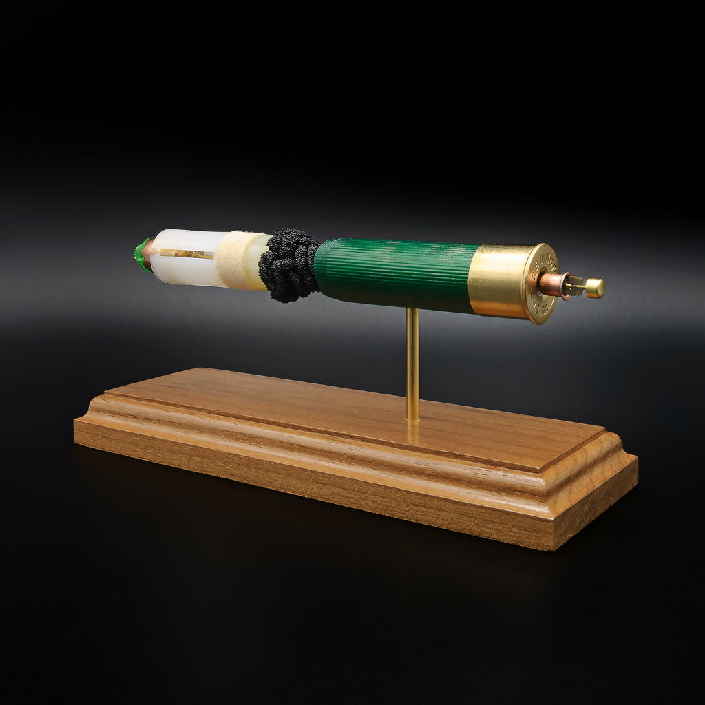 Rear-right perspective of a handcrafted Remington 12 Gauge AccuTip slug display featuring the expanded primer cup, anvil, battery cup, green hull, exposed simulated powder, sabot, and copper projectile with green polymer tip mounted on a walnut wood base.