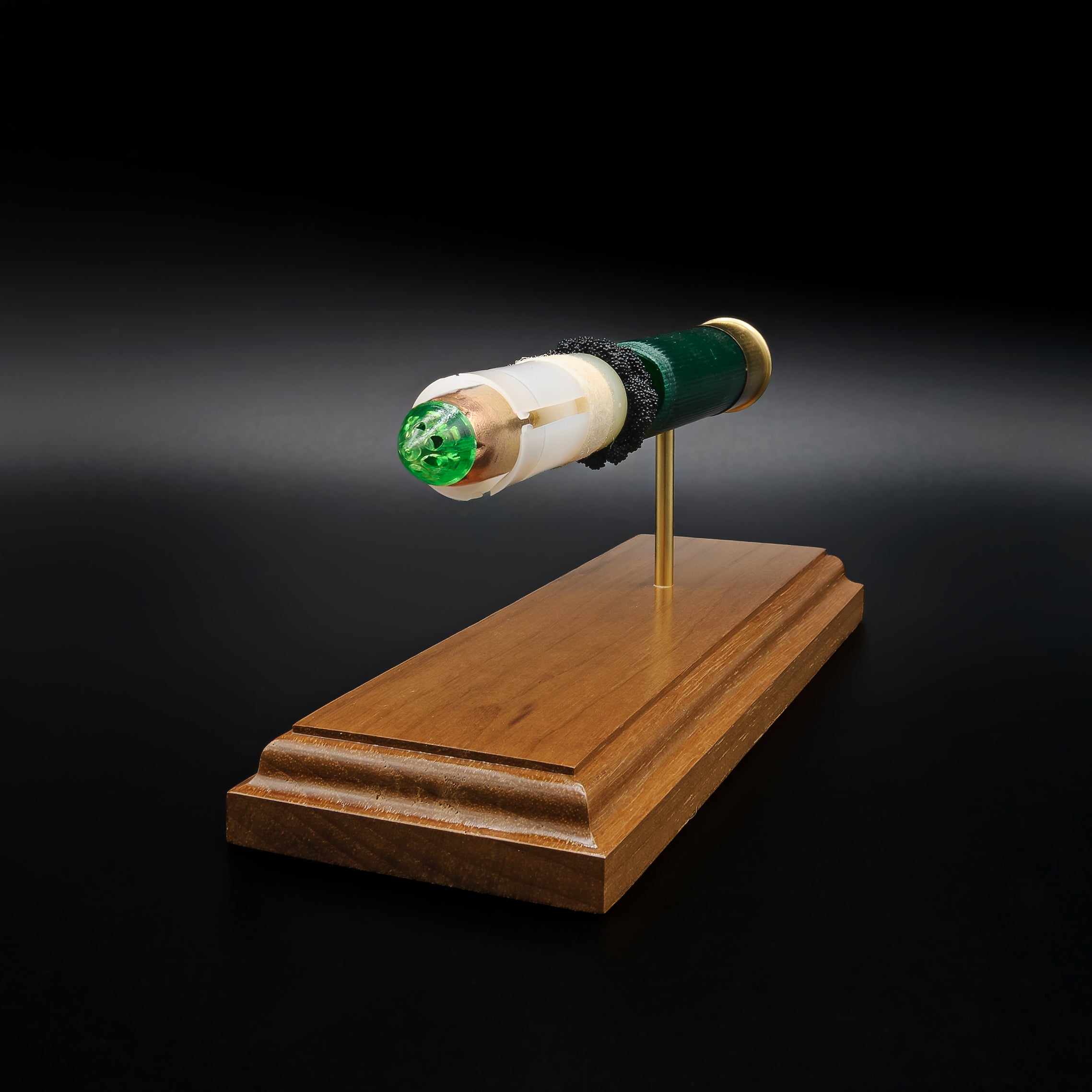 Front-left three-quarter view of an inert Remington 12 Gauge AccuTip display highlighting the sabot surrounding the copper slug with green polymer tip, exposed simulated powder, expanded primer assembly including the primer cup, anvil, and battery cup, and walnut presentation base.