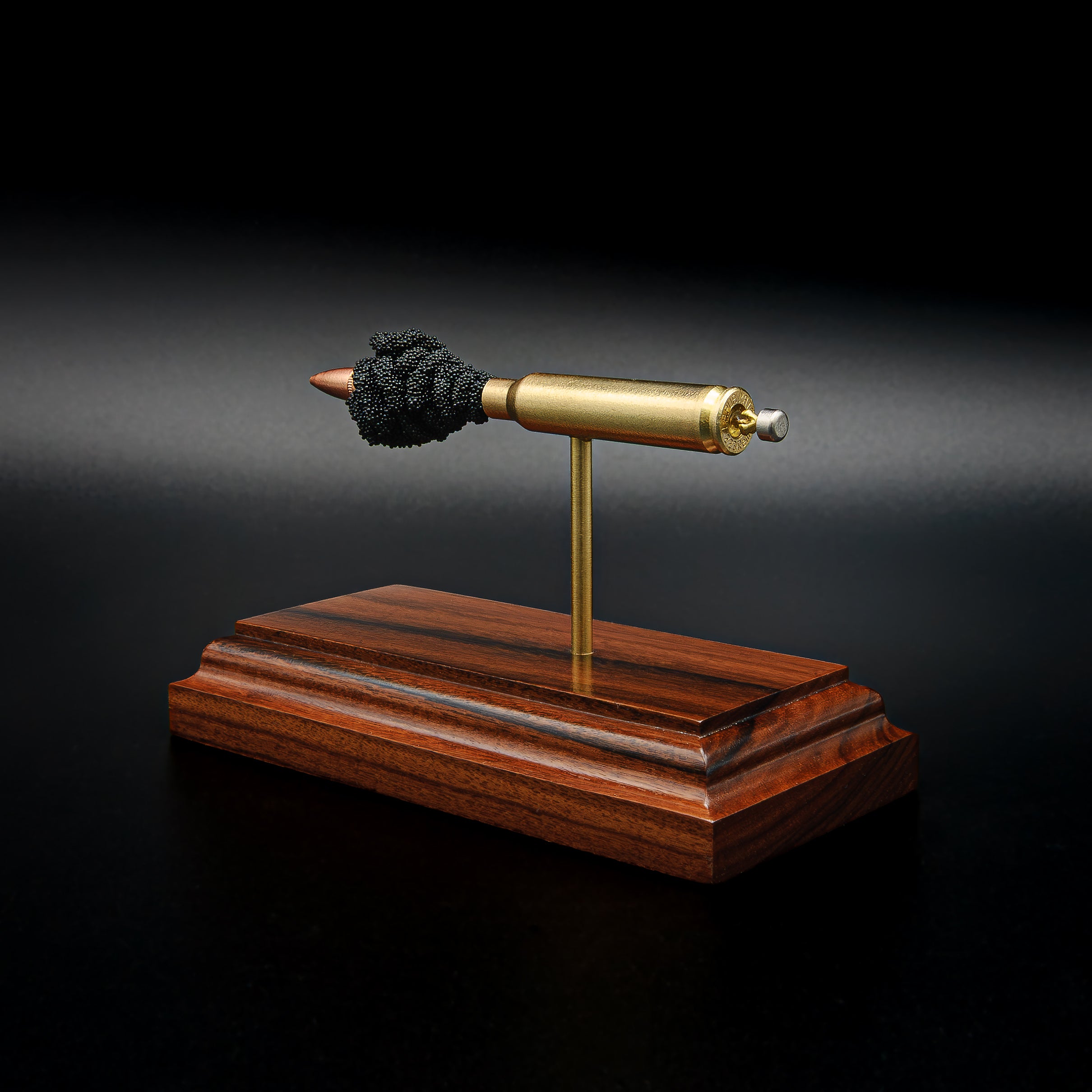 Detailed perspective of a .223 Remington FMJ exploded cartridge display mounted on a handcrafted Bolivian rosewood base, revealing the bullet, brass case, simulated powder, and expanded primer assembly including the primer cup, anvil, and battery cup.