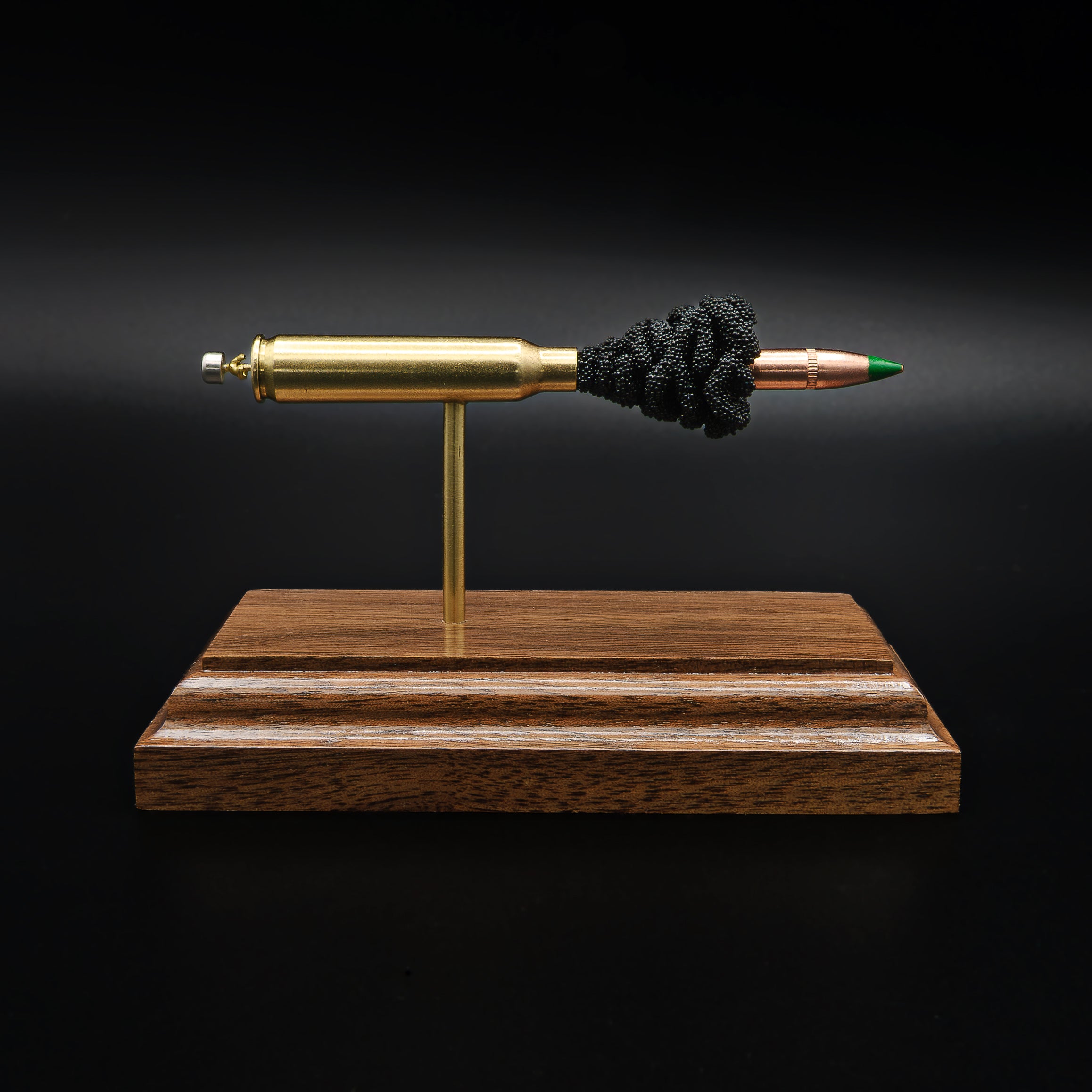 Front view of a 5.56×45mm NATO M855 Green Tip cartridge anatomy display showing the primer cup, anvil, brass case, simulated powder, and steel-core projectile on a walnut wood base.