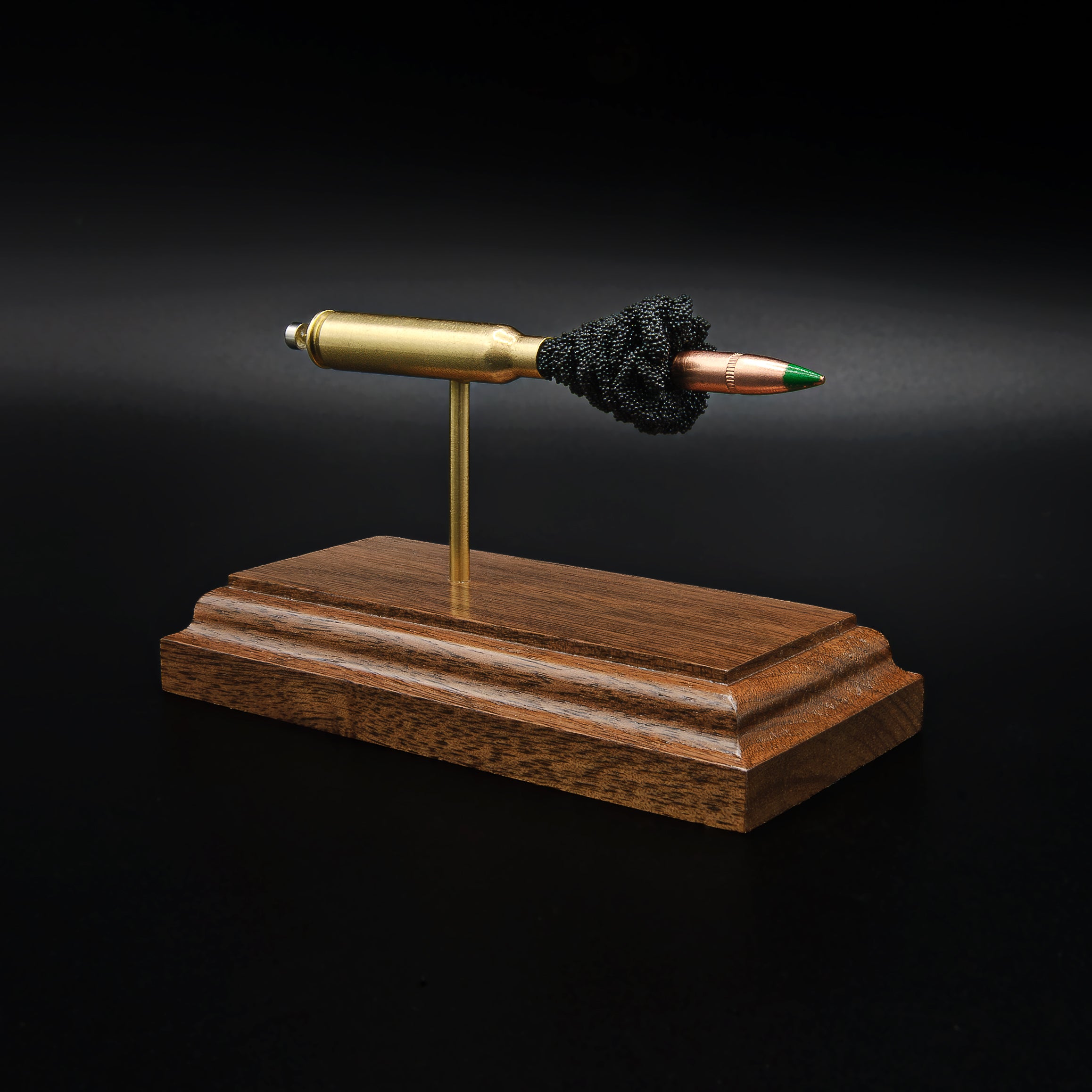 Front left angle of a 5.56×45mm NATO M855 Green Tip cartridge anatomy display highlighting the exposed simulated powder, brass case, and green tip projectile on a walnut base.
