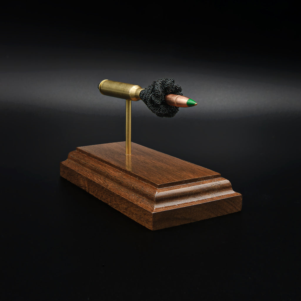 Left side view of a 5.56×45mm NATO M855 Green Tip cartridge anatomy display with the primer cup, anvil, brass case, simulated powder, and projectile visible on a walnut base.