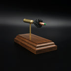 Left side view of a 5.56×45mm NATO M855 Green Tip cartridge anatomy display with the primer cup, anvil, brass case, simulated powder, and projectile visible on a walnut base.