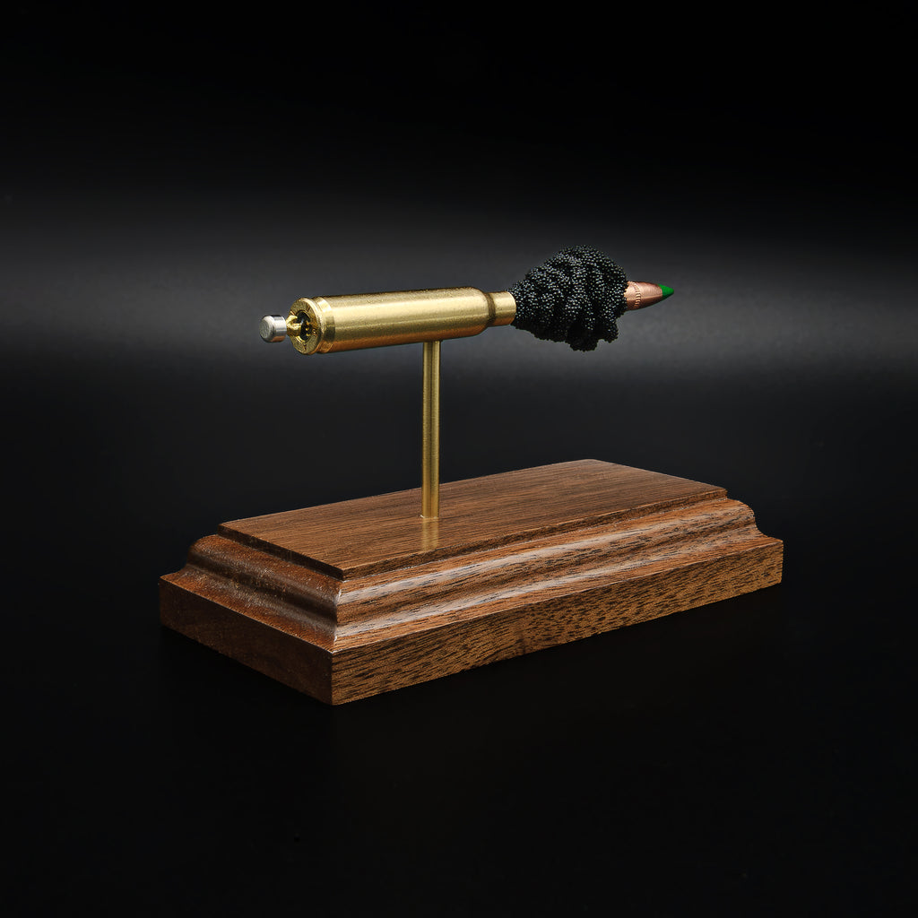 Rear left angle of a 5.56×45mm NATO M855 Green Tip cartridge anatomy display showing the cartridge head, primer assembly, and separated internal components on a walnut base.