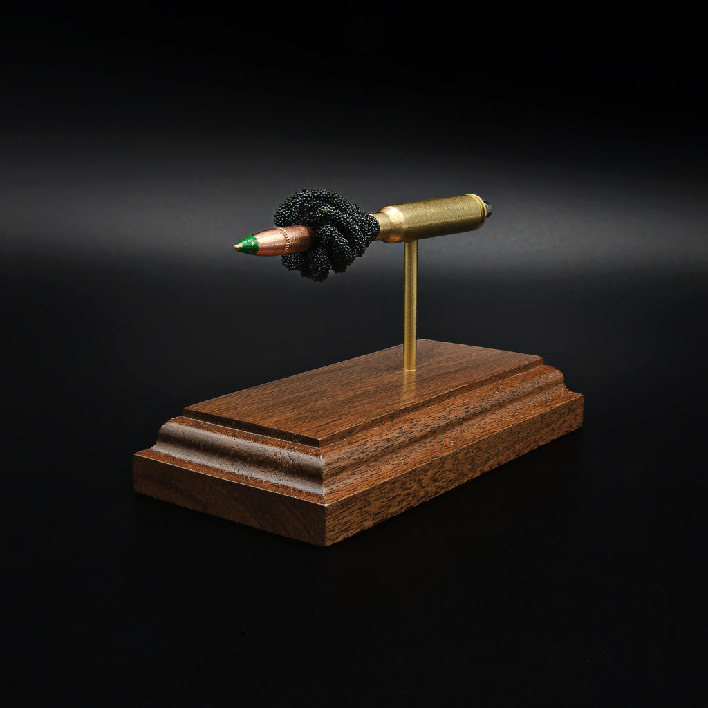 Front left angle of a 5.56×45mm NATO M855 Green Tip cartridge anatomy display with the primer cup, anvil, brass case, simulated powder, and projectile mounted on a walnut base.