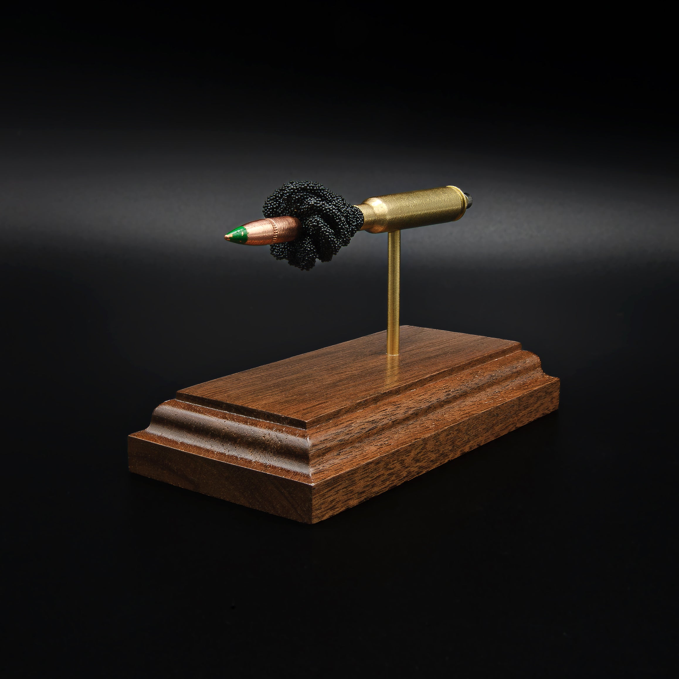 Front left angle of a 5.56×45mm NATO M855 Green Tip cartridge anatomy display with the primer cup, anvil, brass case, simulated powder, and projectile mounted on a walnut base.
