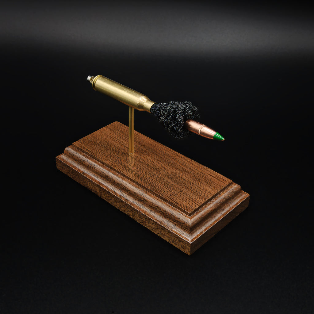 Top front angle of a 5.56×45mm NATO M855 Green Tip cartridge anatomy display showcasing the green tip projectile, simulated powder, brass case, and expanded primer assembly on a walnut base.