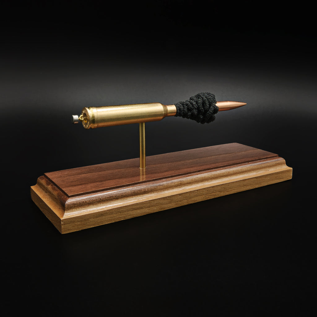 Rear-left three-quarter view of an inert 7mm Remington Magnum display highlighting the cartridge case head, primer assembly, 168 grain Berger VLD projectile, and simulated powder on a walnut presentation base.