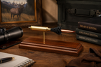 Expanded 7mm PRC Hornady Precision Hunter 180gr ELD-M cartridge display mounted on a polished Bolivian rosewood base, featuring the visible primer cup, anvil, cartridge case, simulated powder, and projectile. Displayed on a rustic wooden desk with a spotting scope, hunting books, notebook, leather gloves, and wildlife artwork in a warmly lit hunting lodge office.