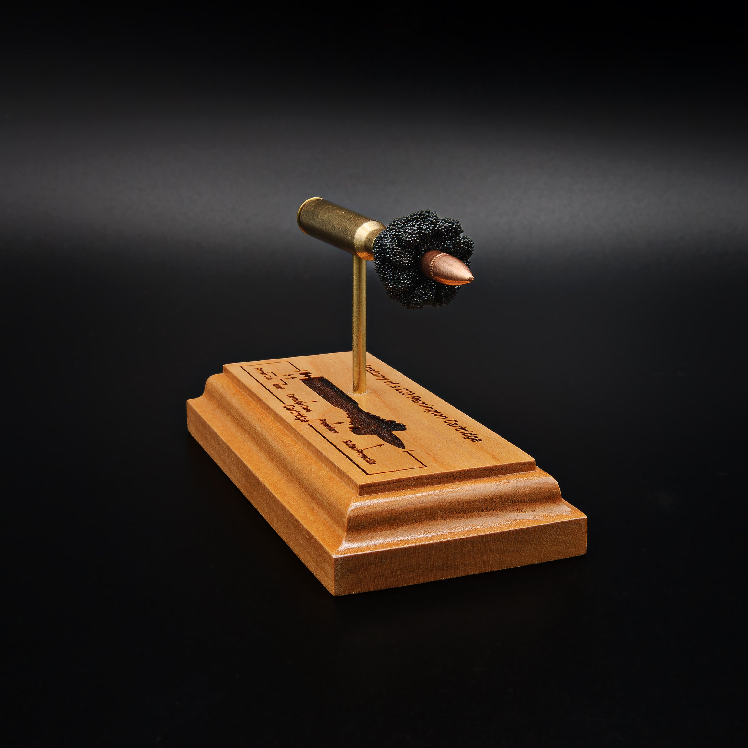 Angled front view of a .223 Remington FMJ cartridge anatomy display featuring the primer cup, anvil, cartridge case, simulated powder, and projectile on a cherry wood base with an engraved anatomy diagram.