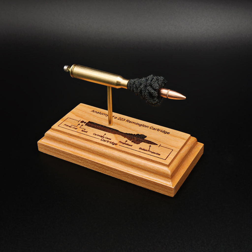 Three-quarter view of a .223 Remington FMJ cartridge anatomy display showing the primer cup, anvil, cartridge case, simulated powder, and projectile on a cherry wood base with an engraved anatomy diagram.
