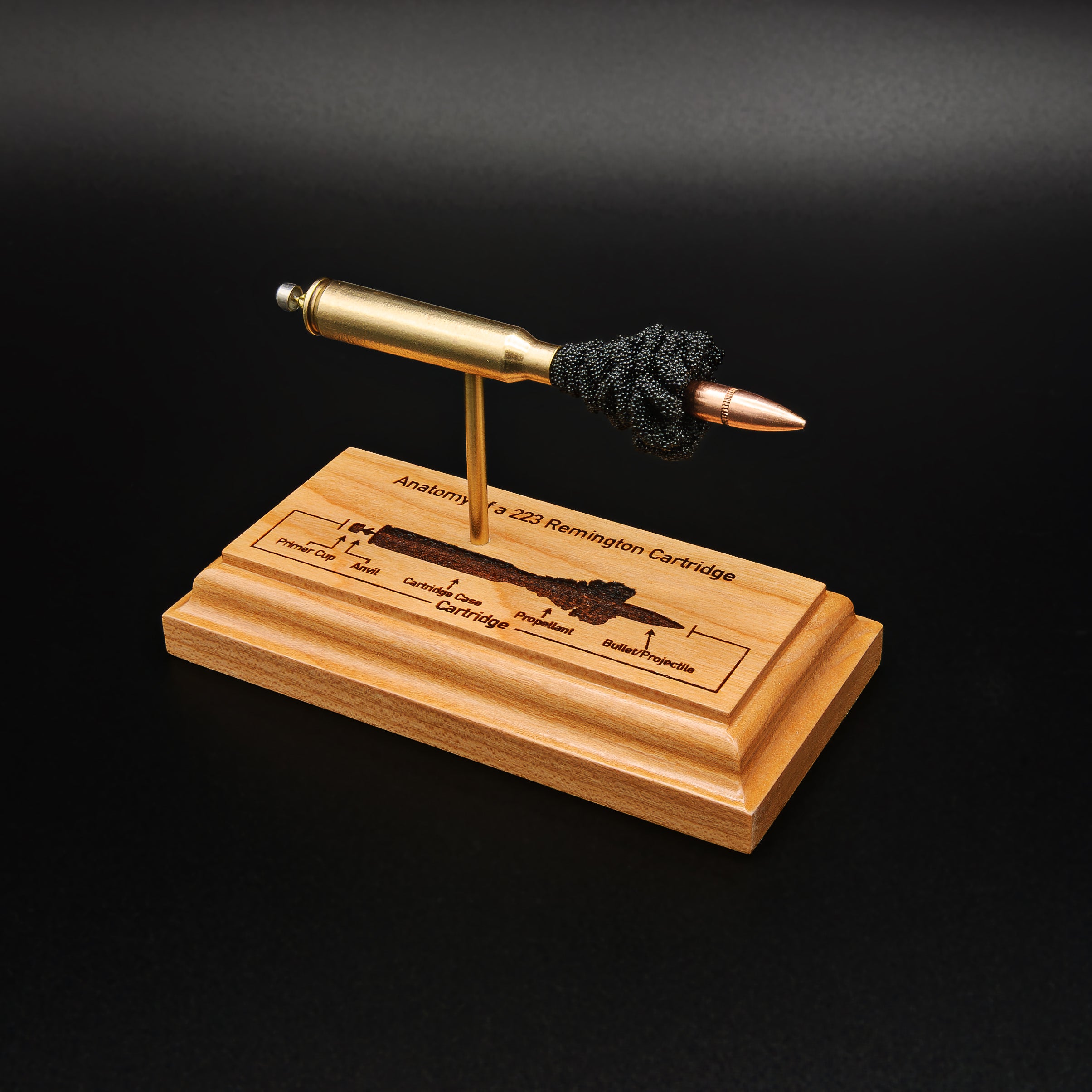 Three-quarter view of a .223 Remington FMJ cartridge anatomy display showing the primer cup, anvil, cartridge case, simulated powder, and projectile on a cherry wood base with an engraved anatomy diagram.