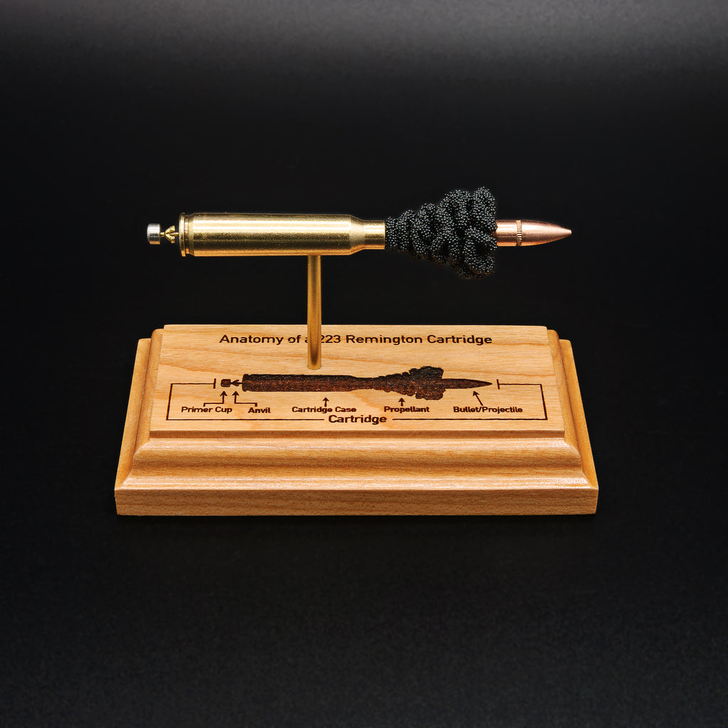 Front view of a .223 Remington FMJ cartridge anatomy display showing the primer cup, anvil, cartridge case, simulated powder, and projectile on a cherry wood base with an engraved anatomy diagram.