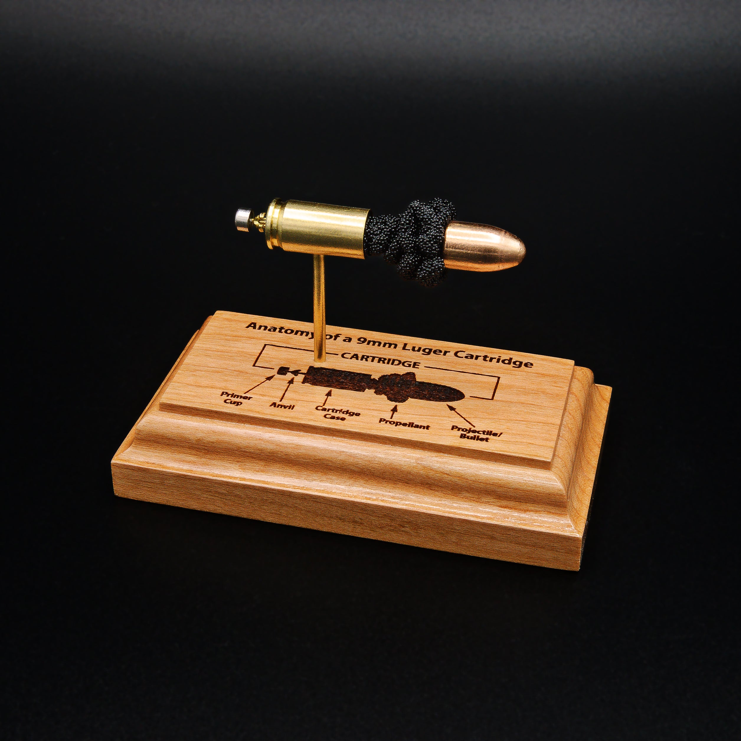 Front angled view of a handcrafted 9mm Luger cartridge anatomy display featuring a 115gr Sierra FMJ bullet with an engraved anatomy diagram showing the primer cup, anvil, cartridge case, powder, and projectile that is mounted on a solid cherry wood base.