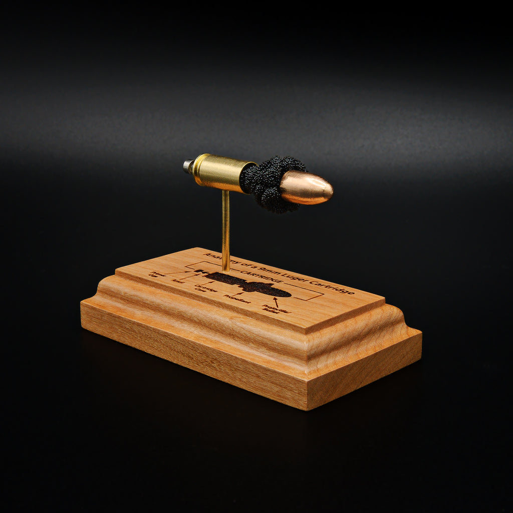 Front right angled view of a handcrafted 9mm Luger cartridge anatomy display featuring a 115gr Sierra FMJ bullet with an engraved anatomy diagram showing the primer cup, anvil, cartridge case, powder, and projectile that is mounted on a solid cherry wood base.