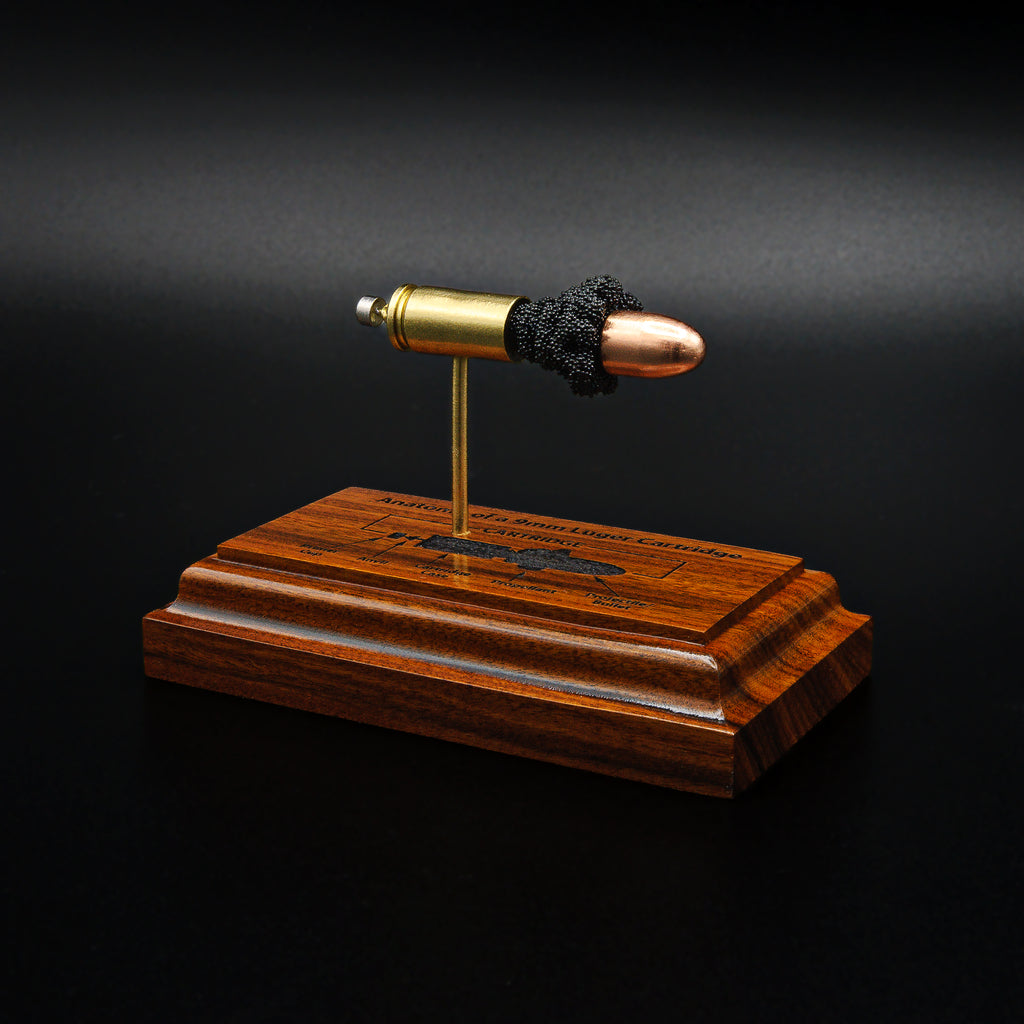 Front angled view of a handcrafted 9mm Luger cartridge anatomy display featuring a 115gr Sierra FMJ bullet with an engraved anatomy diagram showing the primer cup, anvil, cartridge case, powder, and projectile that is mounted on a solid bolivian rosewood base.