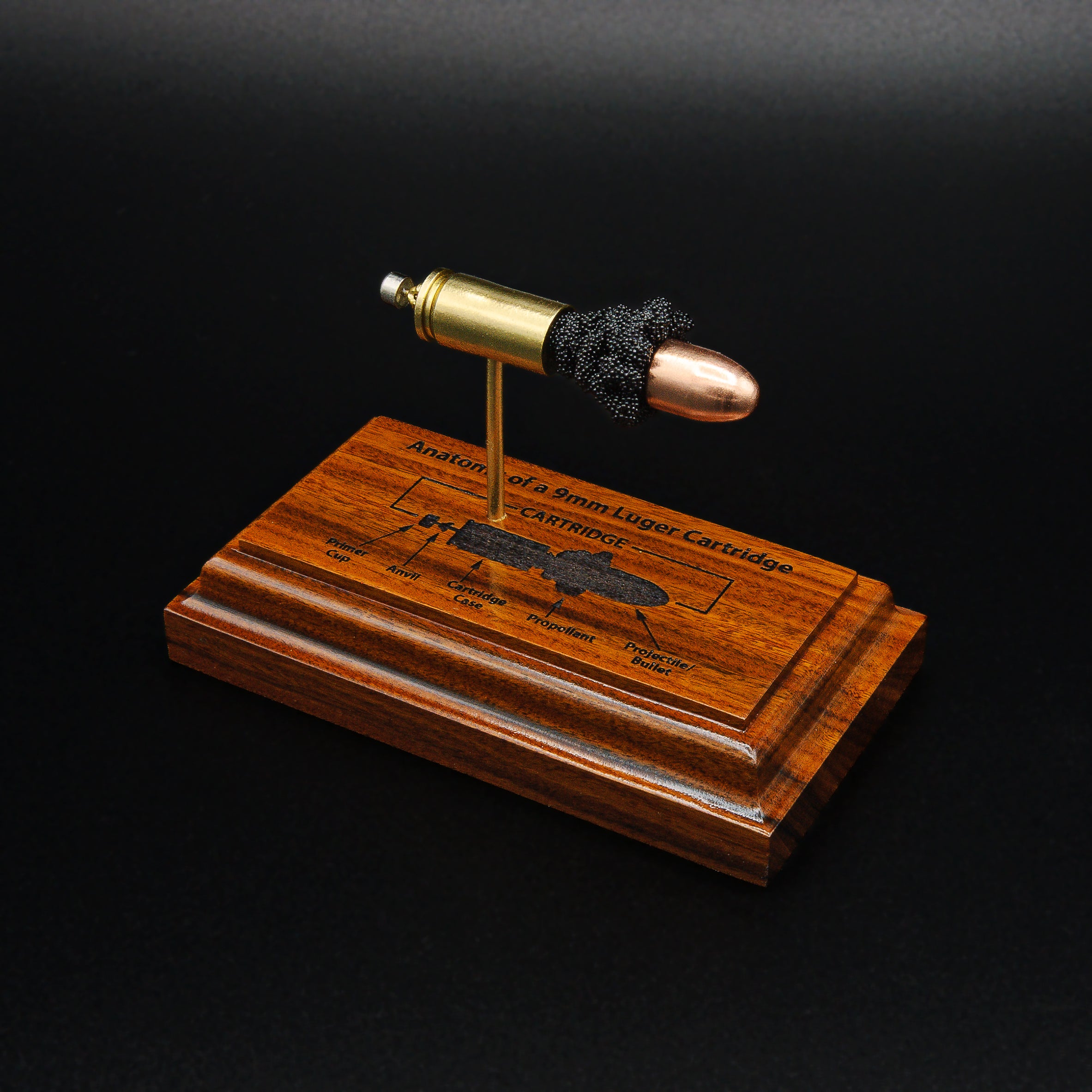 Elevated front right angled view of a handcrafted 9mm Luger cartridge anatomy display featuring a 115gr Sierra FMJ bullet with an engraved anatomy diagram showing the primer cup, anvil, cartridge case, powder, and projectile that is mounted on a solid Bolivian rosewood base.
