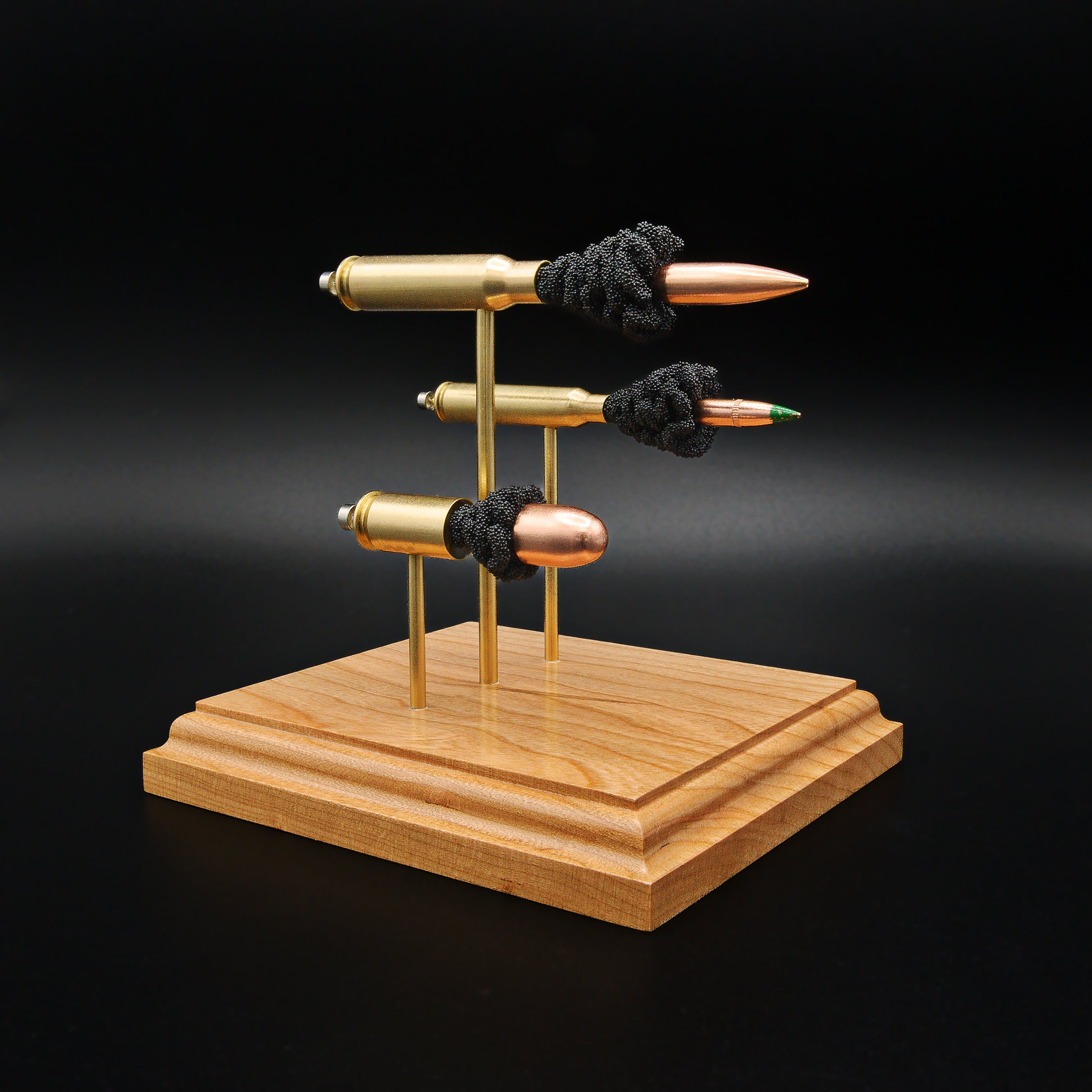 Front left angle of a triple caliber anatomy ammunition display showcasing a .308 Winchester Sierra MatchKing HPBT, 5.56×45mm M855 Green Tip, and .45 Auto Sierra FMJ with simulated powder on a handcrafted cherry wood base.