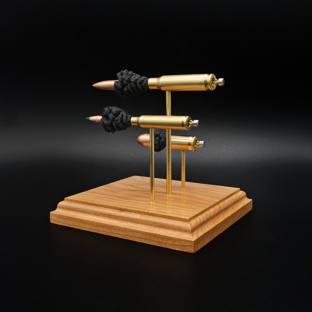 Rear angle of a triple caliber exploded cartridge display showing the brass cases, expanded primer assemblies, and bullet components of a .308 Winchester, 5.56×45mm M855 Green Tip, and .45 Auto on a premium cherry wood base.