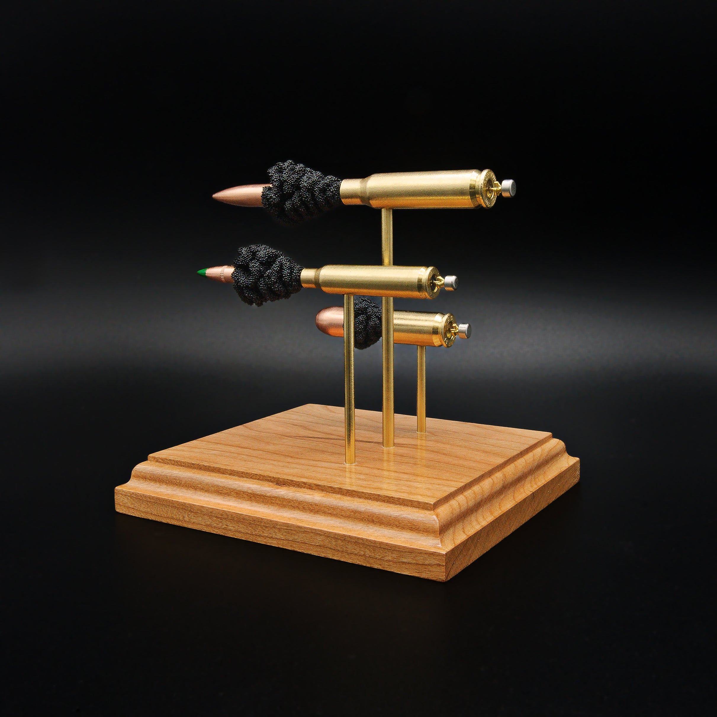 Rear angle of a triple caliber exploded cartridge display showing the brass cases, expanded primer assemblies, and bullet components of a .308 Winchester, 5.56×45mm M855 Green Tip, and .45 Auto on a premium cherry wood base.